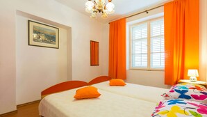 Une chambre d’hôtel avec deux lits, des rideaux orange, un lustre, un tableau encadré et une fenêtre avec des stores.