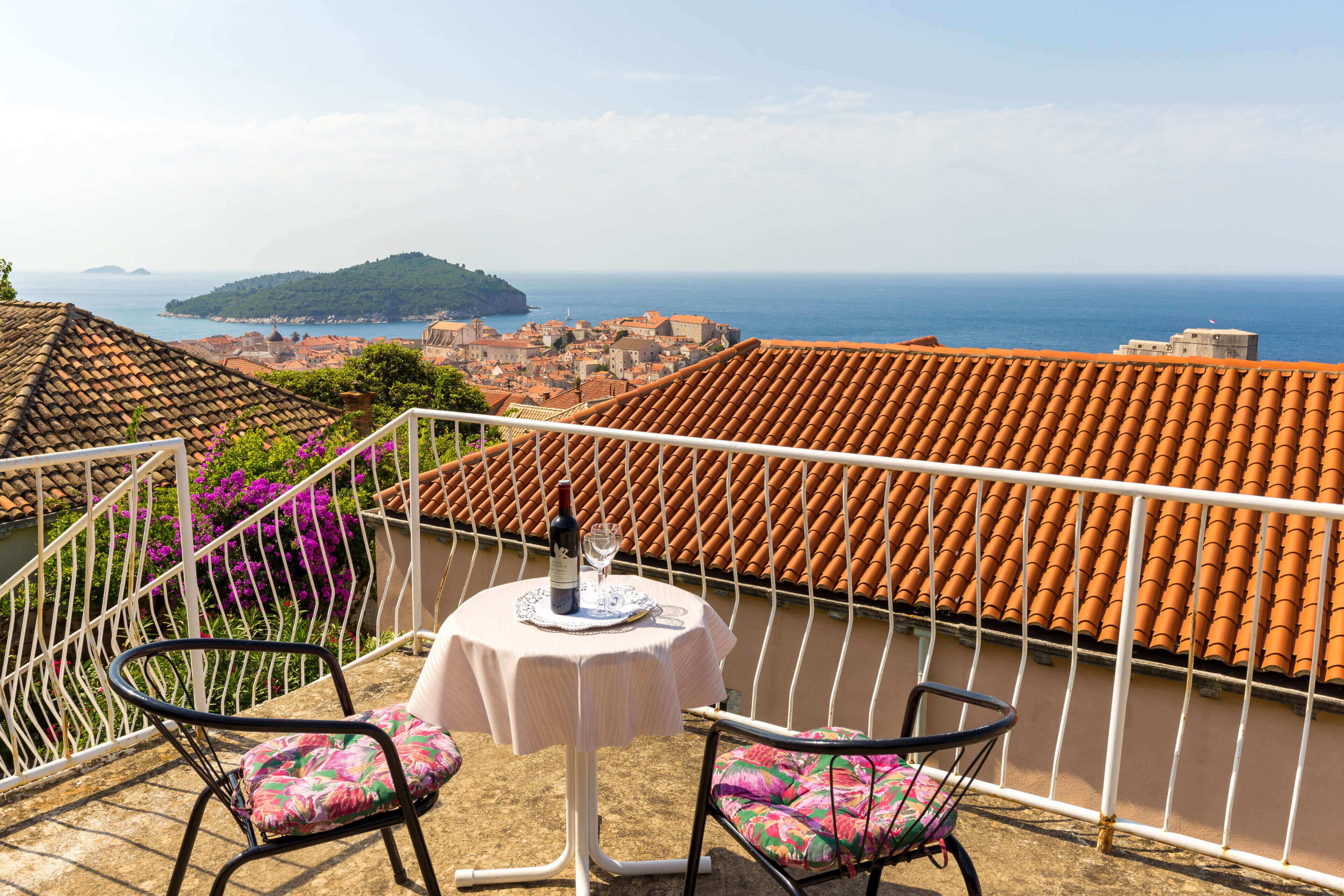 A balcony with a table and chairs, overlooking a coastal town and the sea.