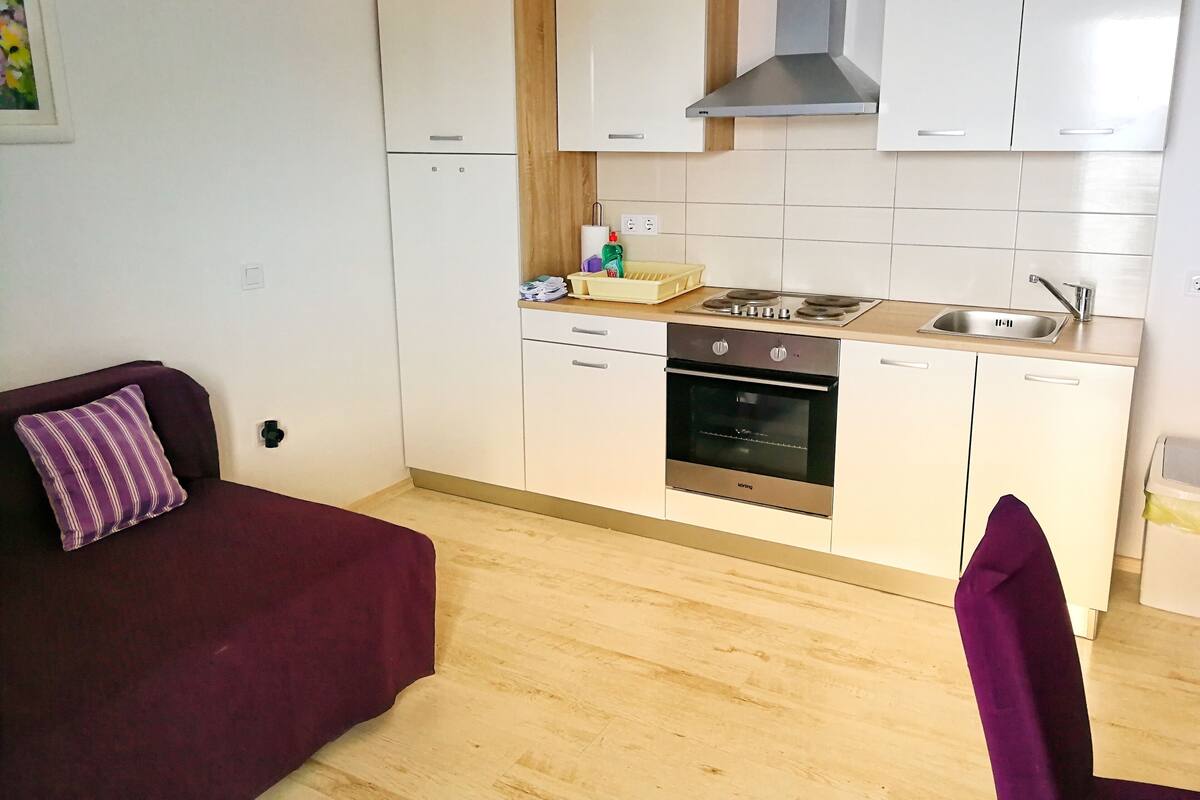 A compact kitchen with a stove, oven, and sink. There is a purple chair and a small table with a vase.
