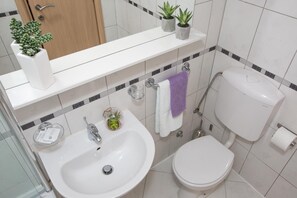 A modern bathroom with a white sink, toilet, and a mirror above the sink. There are two potted plants on the sink's edge, and a towel rack with a purple towel hanging. - Apartments Koncul (Dubrovnik)