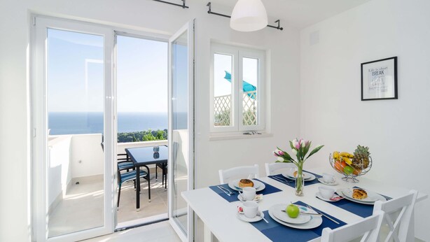 A dining area with a table set for two, a view of the sea through a sliding glass door, and a framed quote on the wall. - Cin & Co Central Apartments (Dubrovnik)