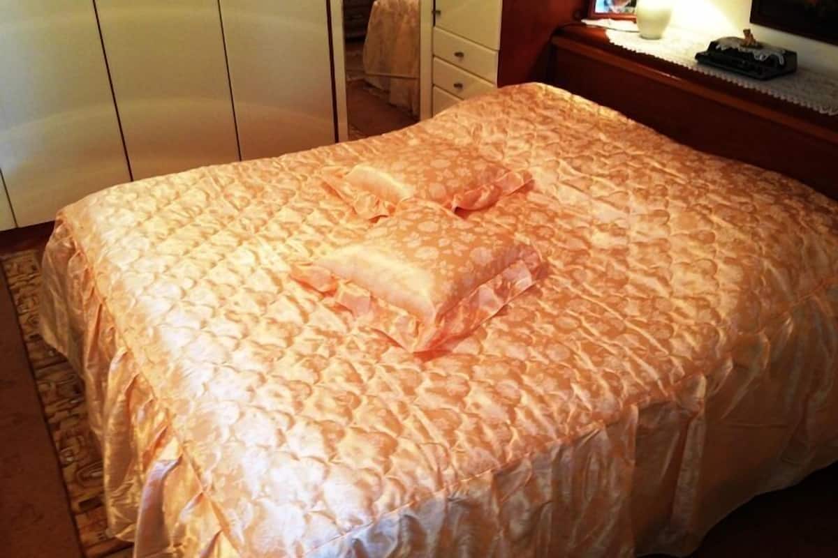 A neatly made bed with a patterned bedspread and pillows, a wooden nightstand with a lamp, and a wardrobe in the background.