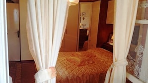Una habitación con una cama cubierta por una colcha estampada, cortinas sujetas en las esquinas, un armario de madera y una mesita de noche con una lámpara.