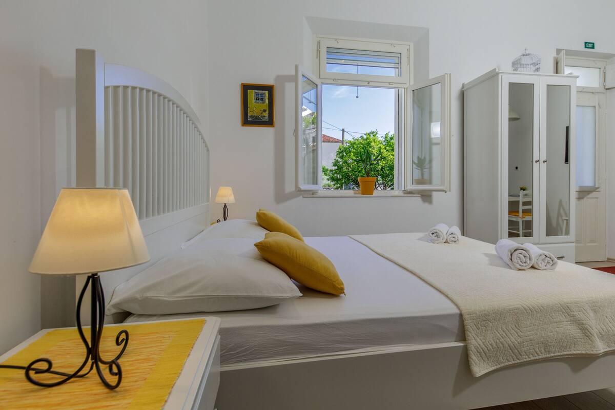 Quarto bem arrumado com uma cama, luminária de cabeceira, janela com vista para o verde e um guarda-roupa.