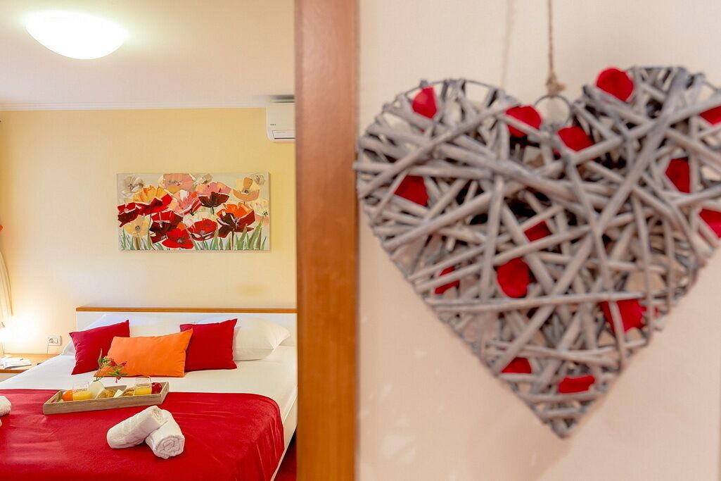 A hotel room with a bed, red and white pillows, a tray with breakfast items, and a painting of flowers on the wall.