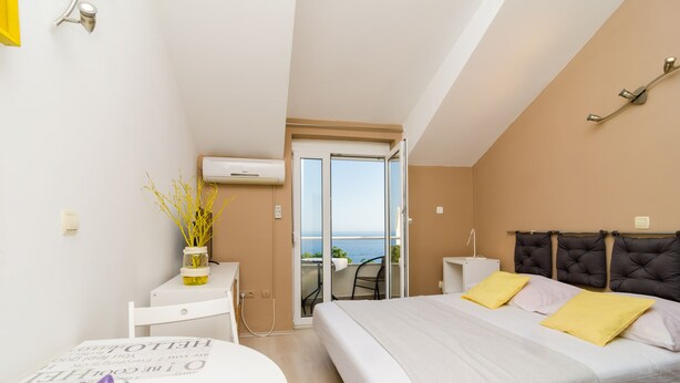A modern hotel room with a bed, a small table with a vase, a chair, and a view of the sea through a sliding door. - Maraština Suites (Dubrovnik)