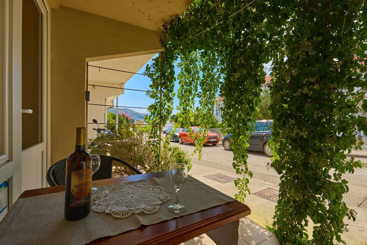 A table with a bottle and a glass on a doily, a chair, and a view of parked cars and buildings through a doorway.
