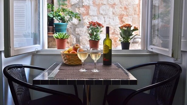 A table with a bottle of wine, two glasses, a bowl of fruit, and potted plants on a windowsill. - Studio Inn (Dubrovnik)