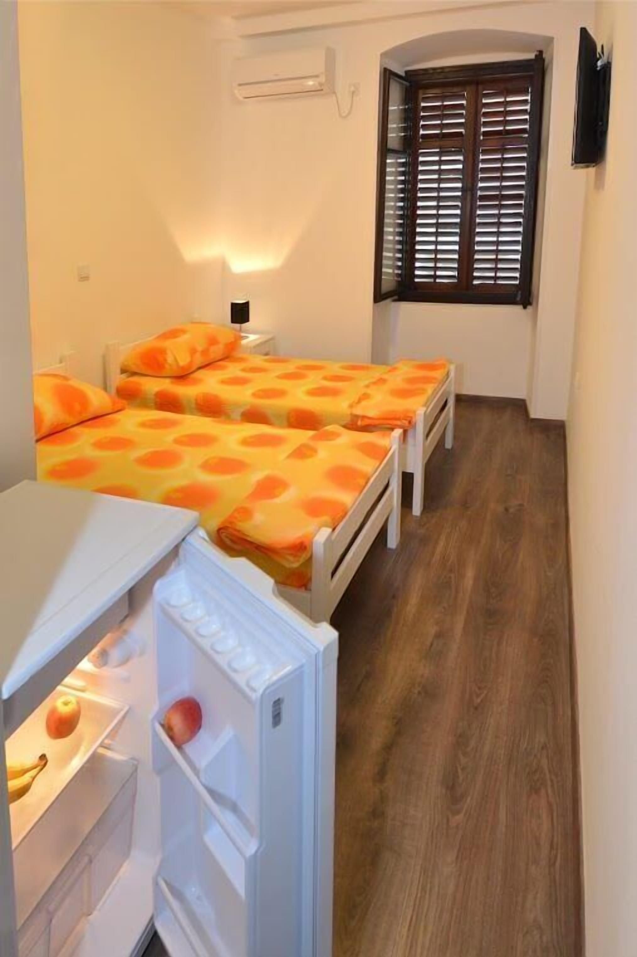A compact room with two beds, a small white table, a refrigerator, and a window with wooden shutters.
