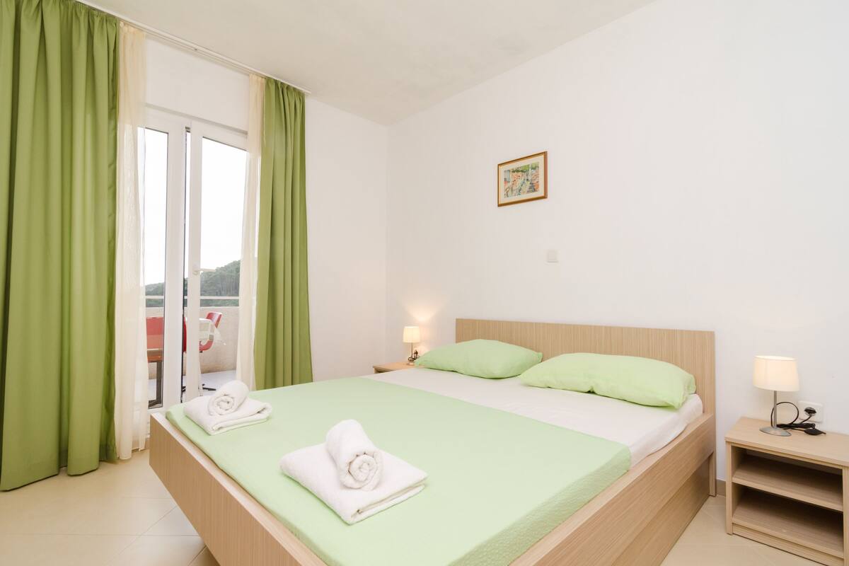 A hotel room with a bed, green curtains, a nightstand, and a view through a glass door.