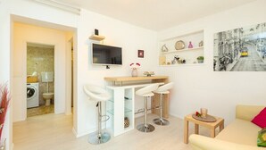 Un salon moderne avec un canapé, une petite table et un coin bar avec des tabourets. Un téléviseur est fixé au mur, et un tableau encadré est accroché au-dessus du bar.