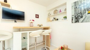 Une cuisine moderne équipée d’un téléviseur à écran plat, de deux tabourets de bar blancs, d’un plan de travail en bois et d’une étagère où sont disposés des objets décoratifs.