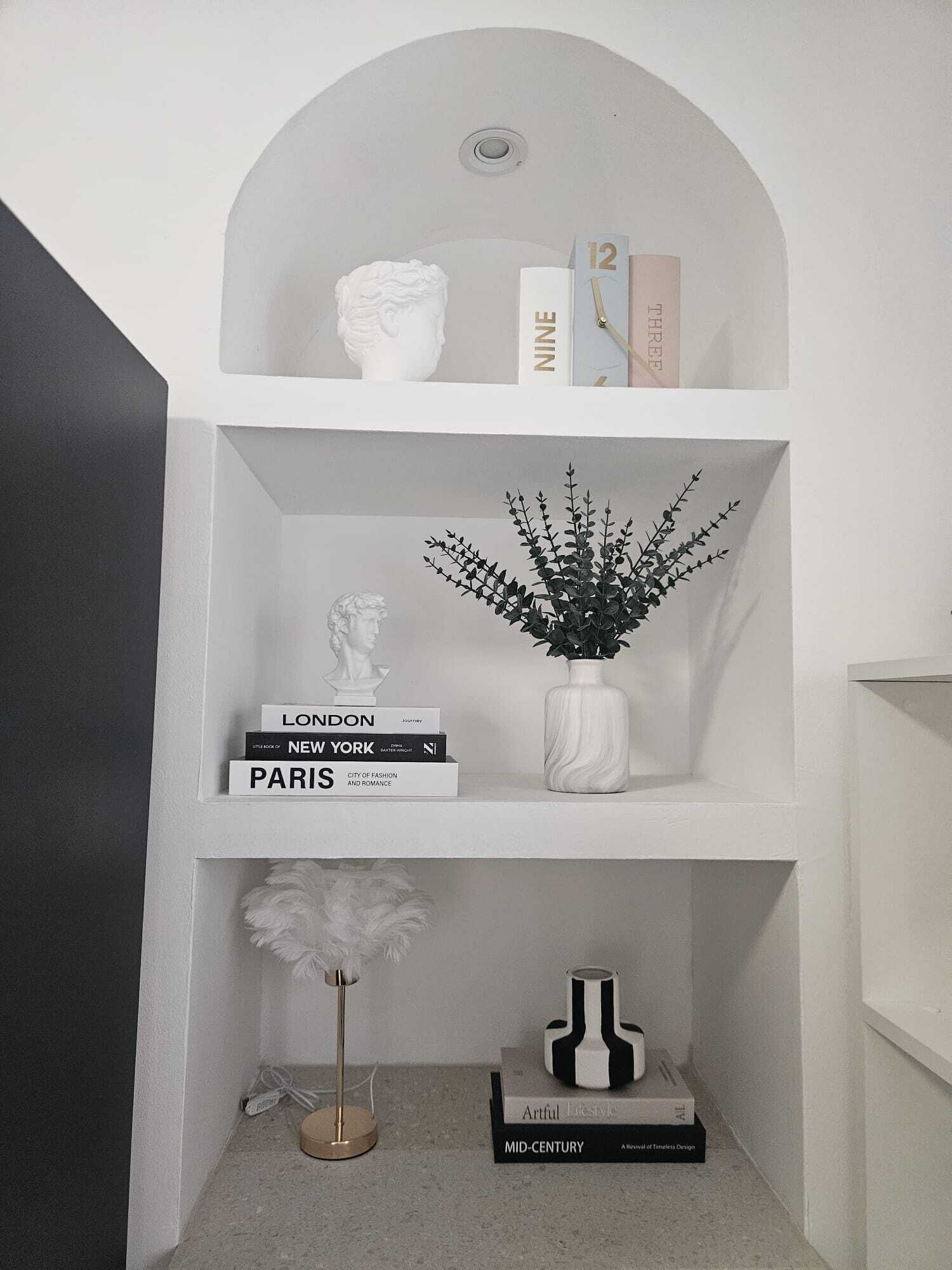 Un interior en tonos blancos con una estantería llena de libros, un jarrón con una planta y un busto.
