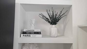 Un interior en tonos blancos con una estantería llena de libros, un jarrón con una planta y un busto.