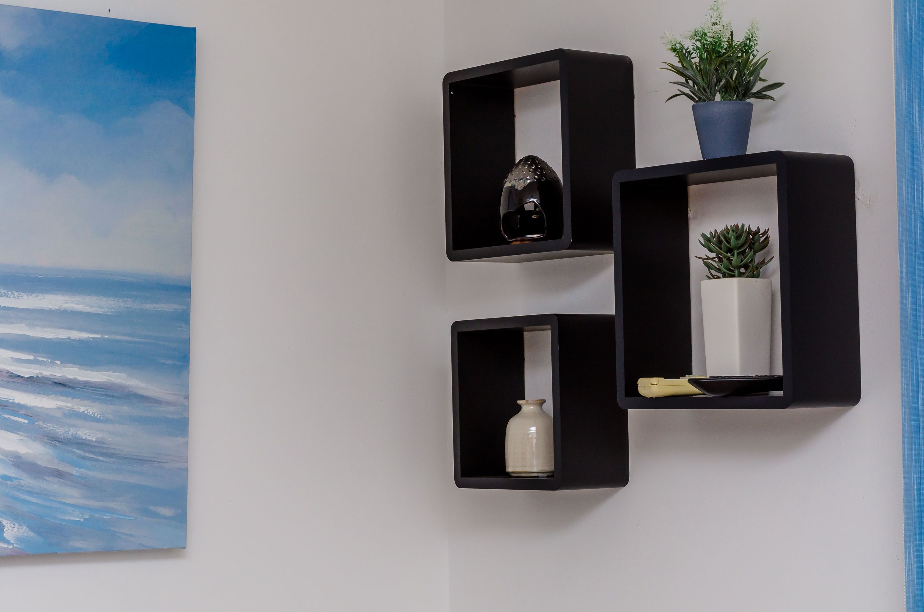Tres estantes negros fijos a la pared, con varios objetos como una planta, un florero y un par de tijeras.