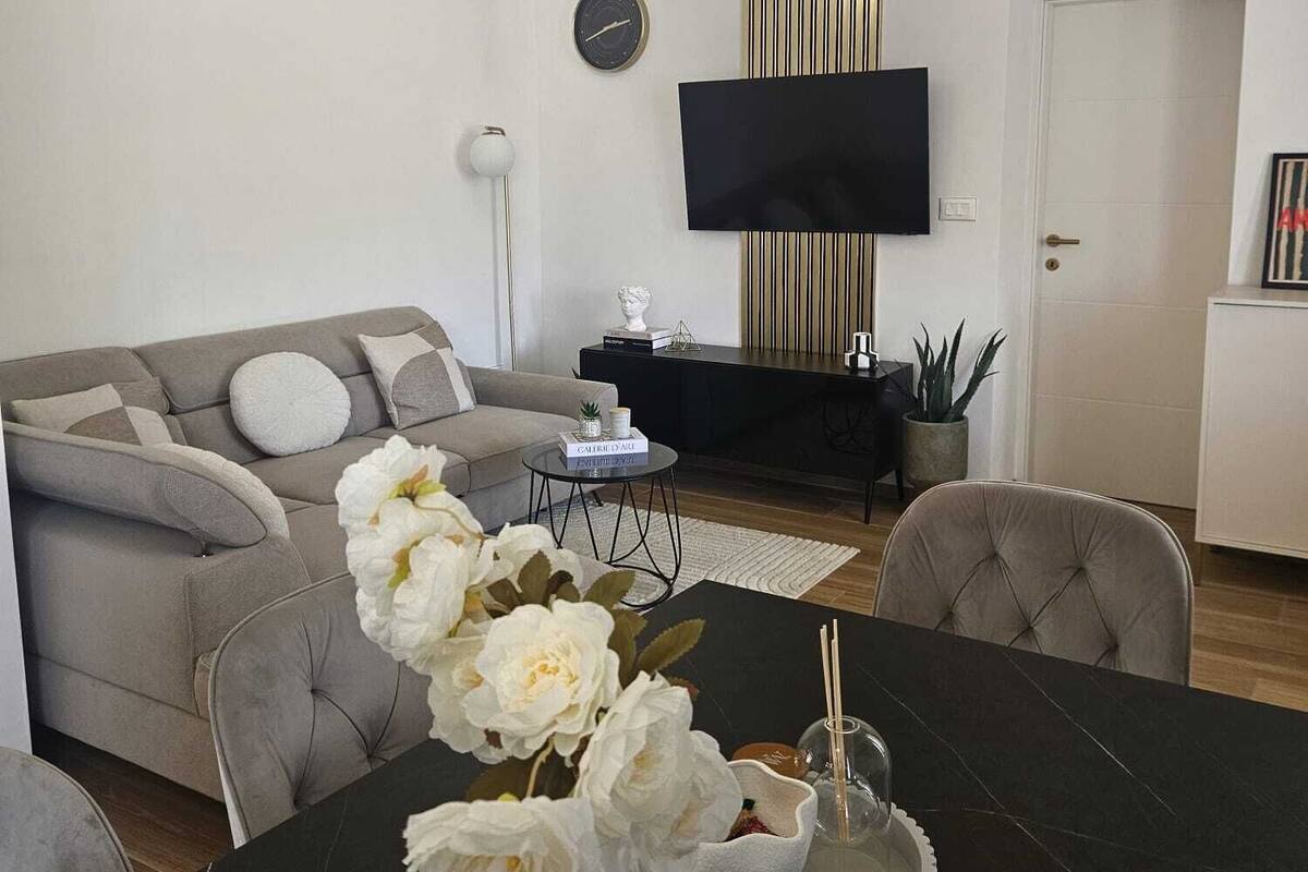 A modern living room with a sofa, a dining table with a vase of flowers, a flat-screen TV mounted on the wall, and a decorative wall panel.