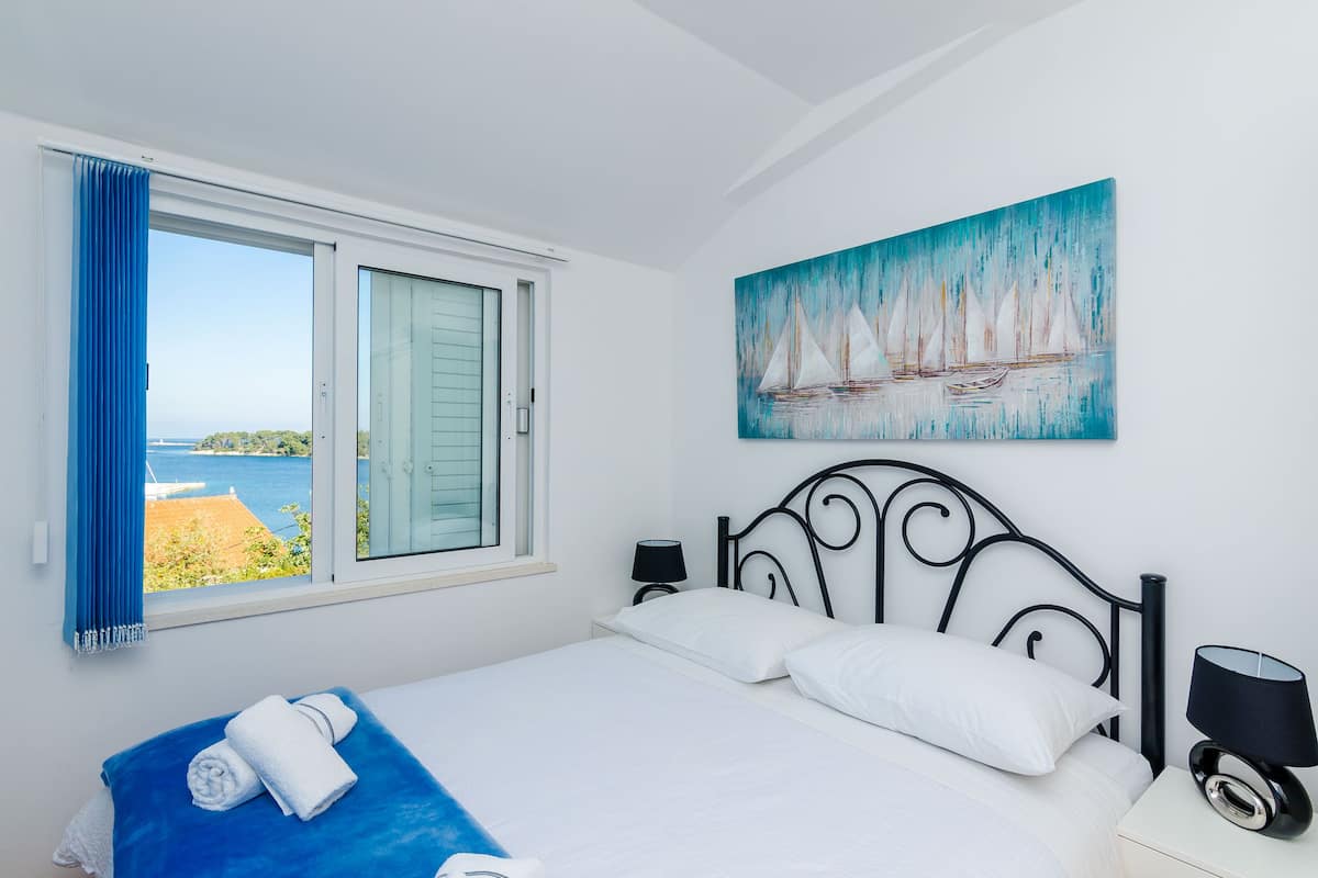 A bedroom with a bed, a window with curtains, a painting of sailboats, and a nightstand with a lamp.