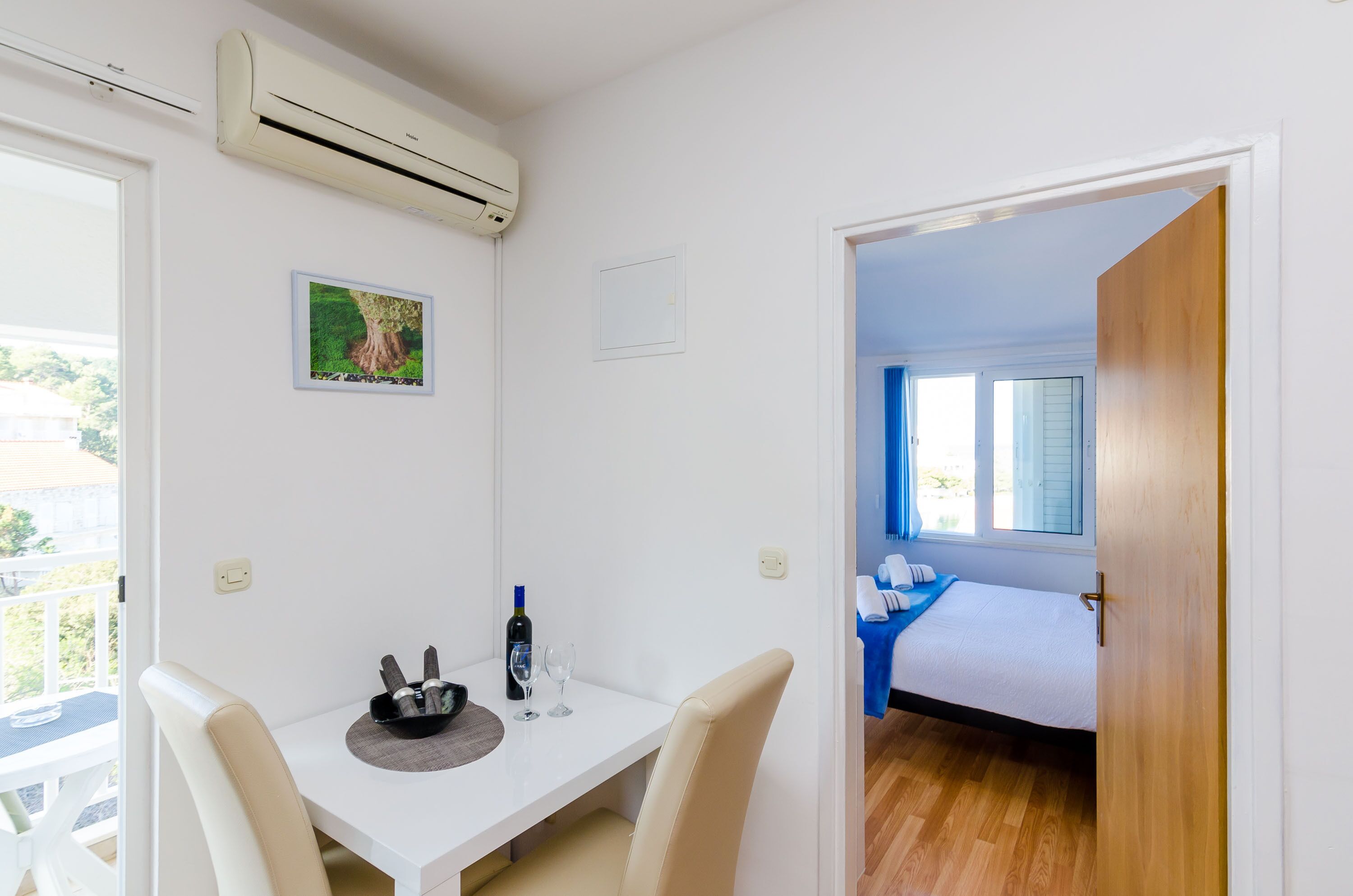 A hotel room with a bed, a small table with a bottle and glasses, an air conditioner, and a window with blinds.