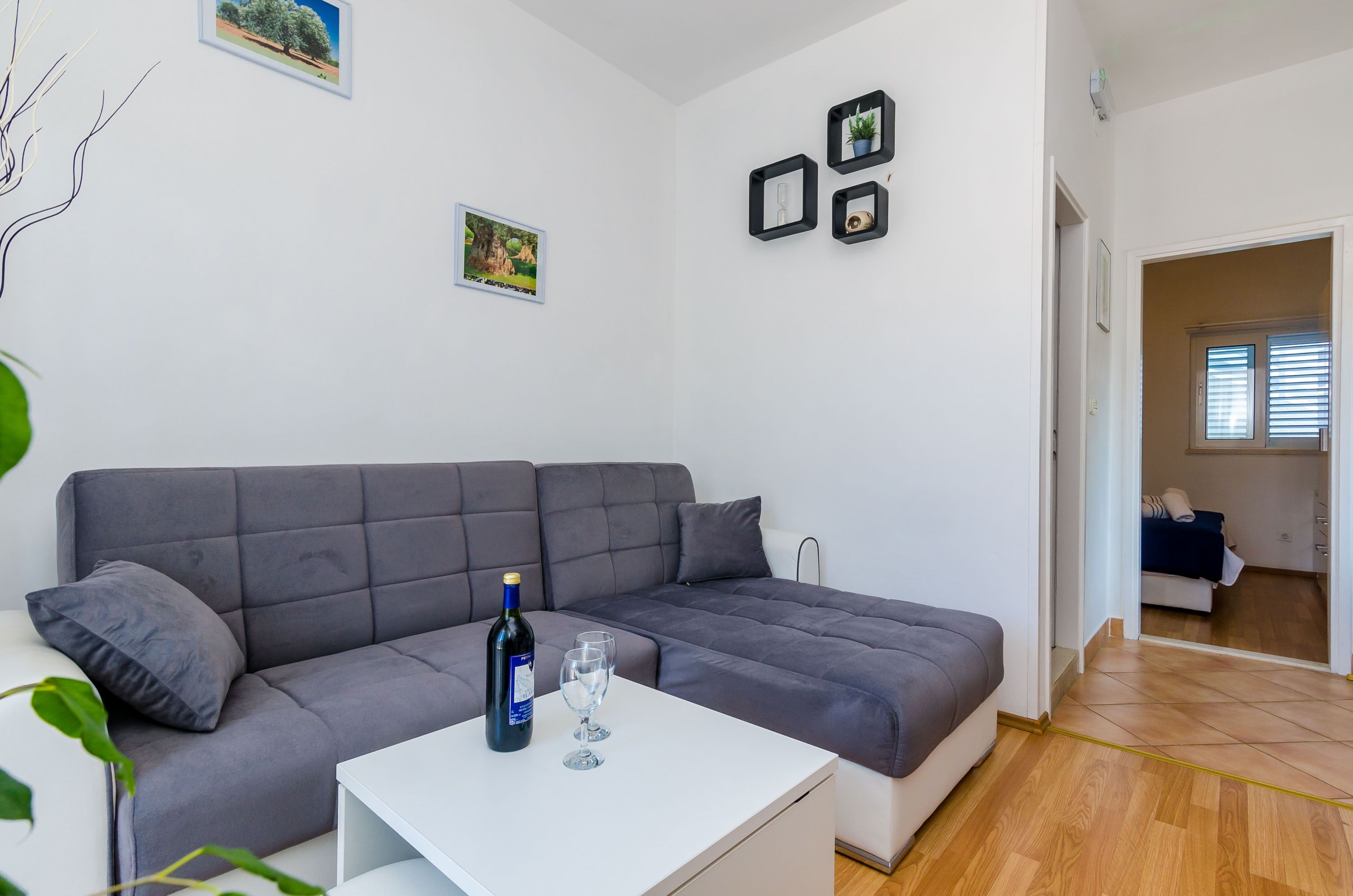Una sala de estar moderna con un sofá gris, una mesa de centro blanca y una botella de vino. Hay cuadros en la pared y una planta a la izquierda.