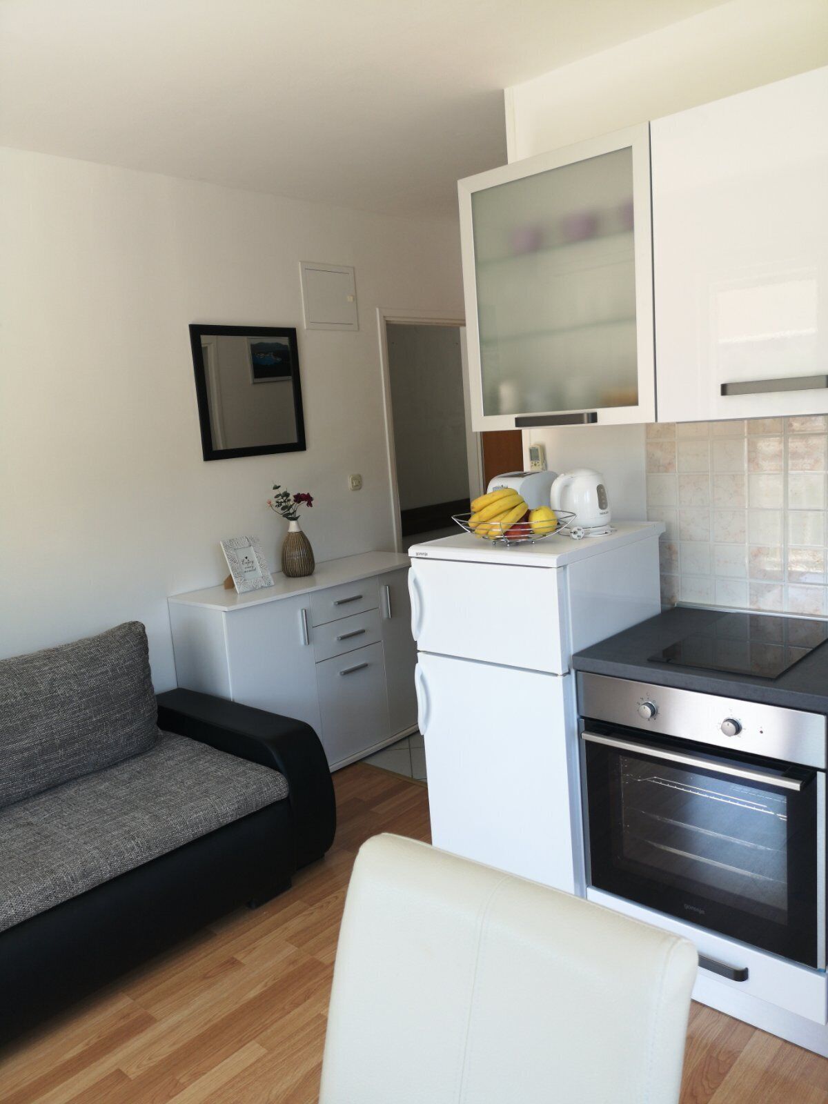 A compact kitchenette with a built-in oven, a small refrigerator, and a microwave. There is a grey sofa and a white chair in the living area.