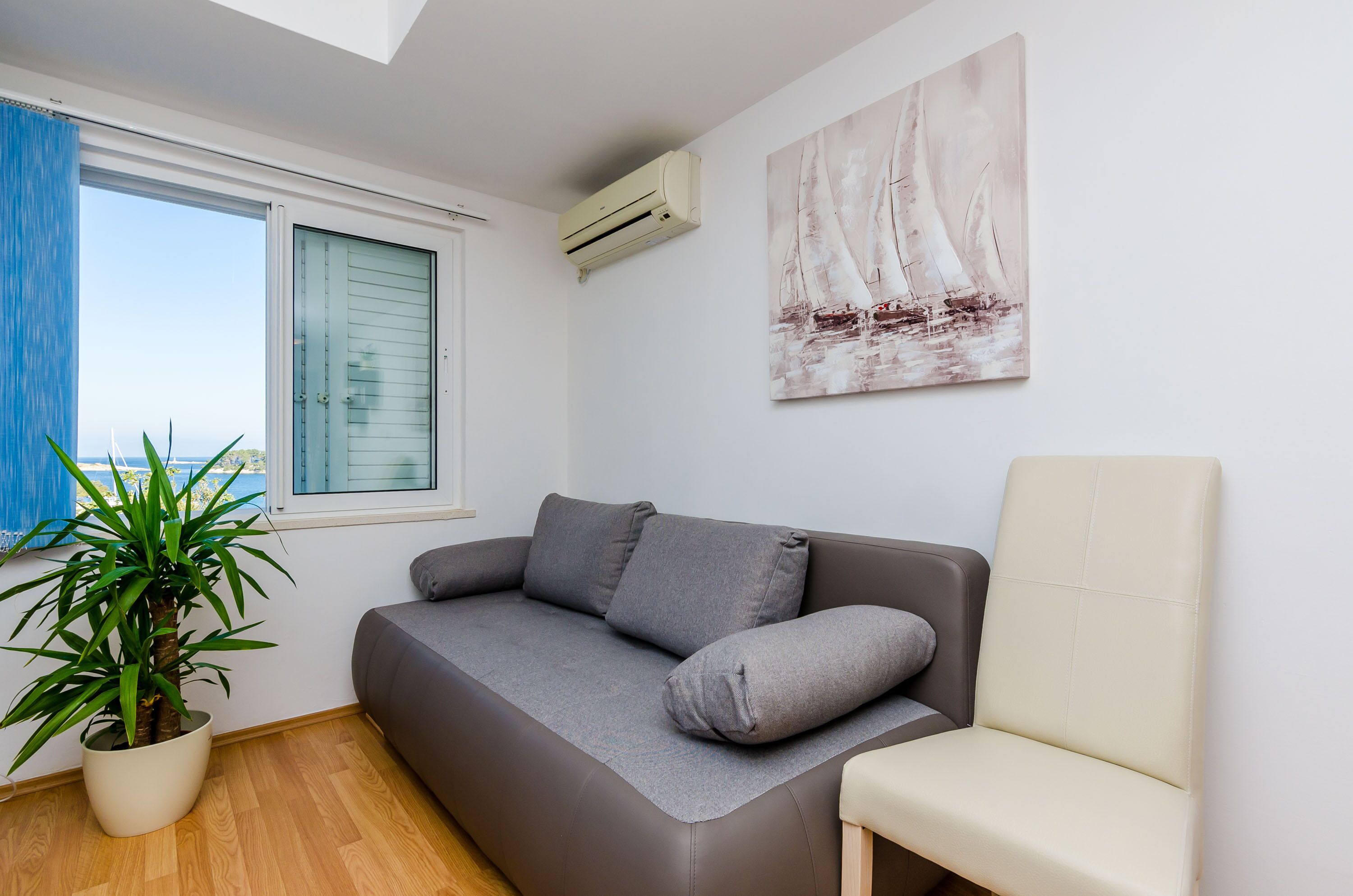 A modern living room with a grey sofa, a white armchair, a large potted plant, and a sailboat painting on the wall.