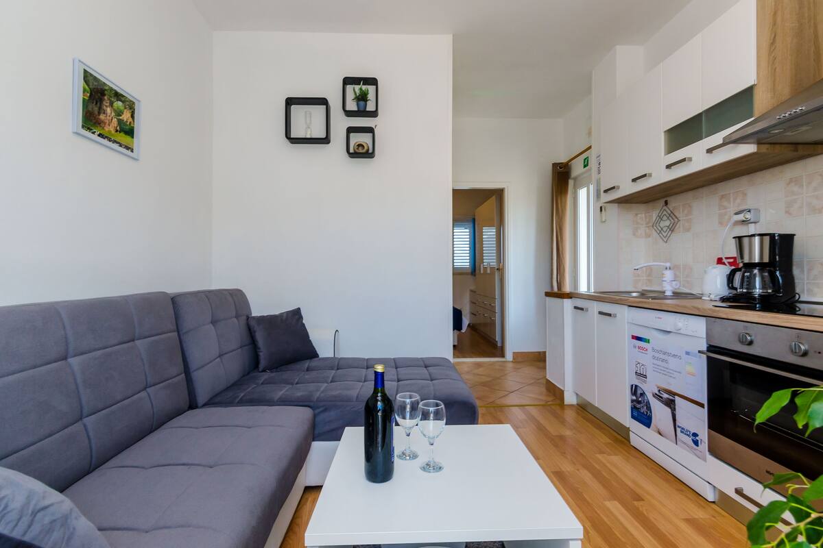 A modern living room with a grey sofa, a white coffee table with a bottle and glasses, and a kitchen with white cabinets and a dishwasher.