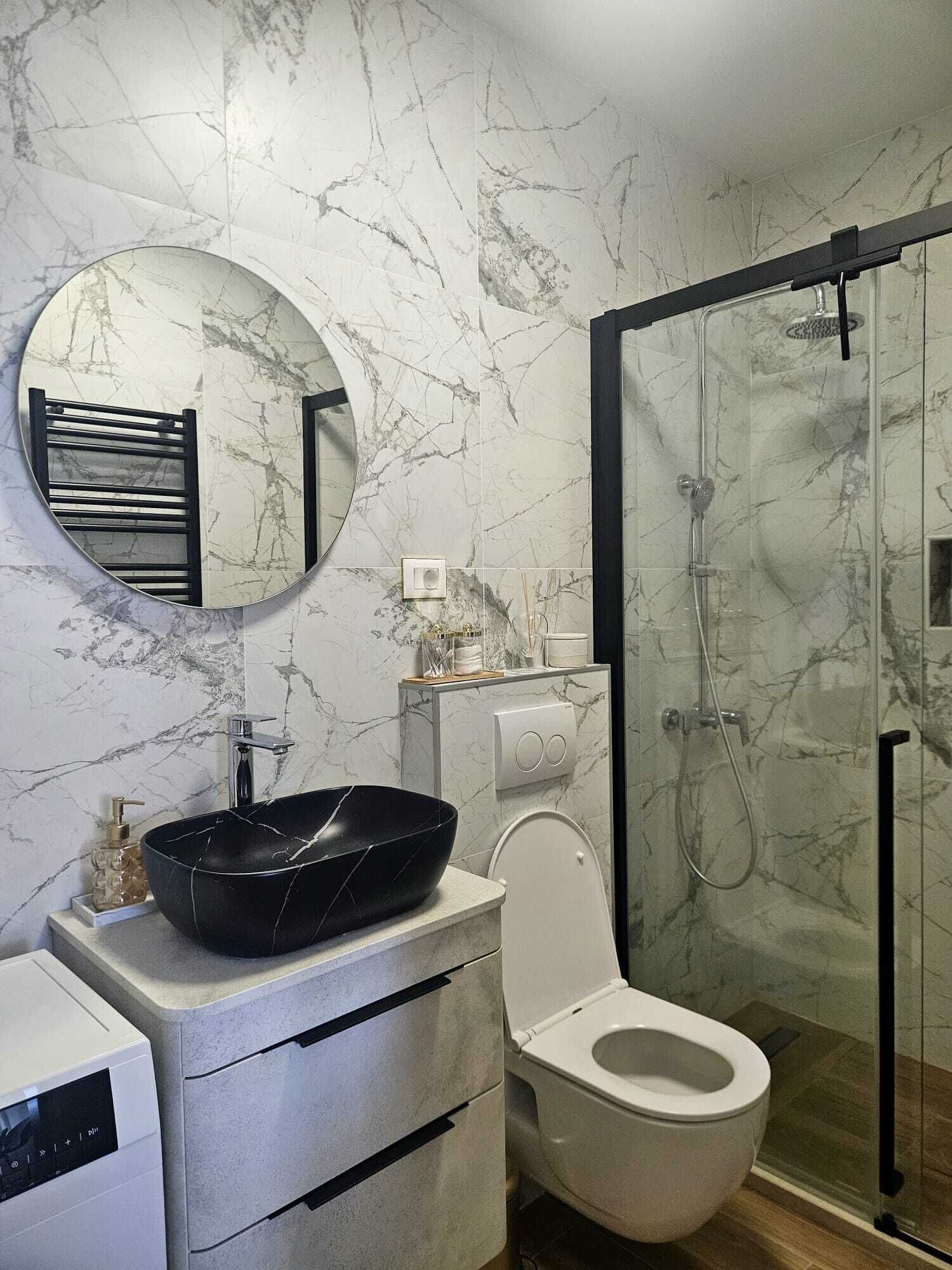 Een moderne badkamer met een marmeren spatwand, een ronde spiegel, een zwart wastafelbakje, een toilet en een douche met glazen wanden.