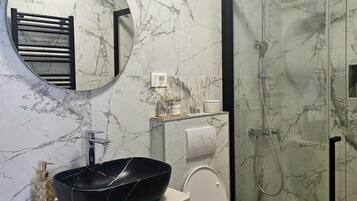 Een moderne badkamer met een marmeren spatwand, een ronde spiegel, een zwart wastafelbakje, een toilet en een douche met glazen wanden.