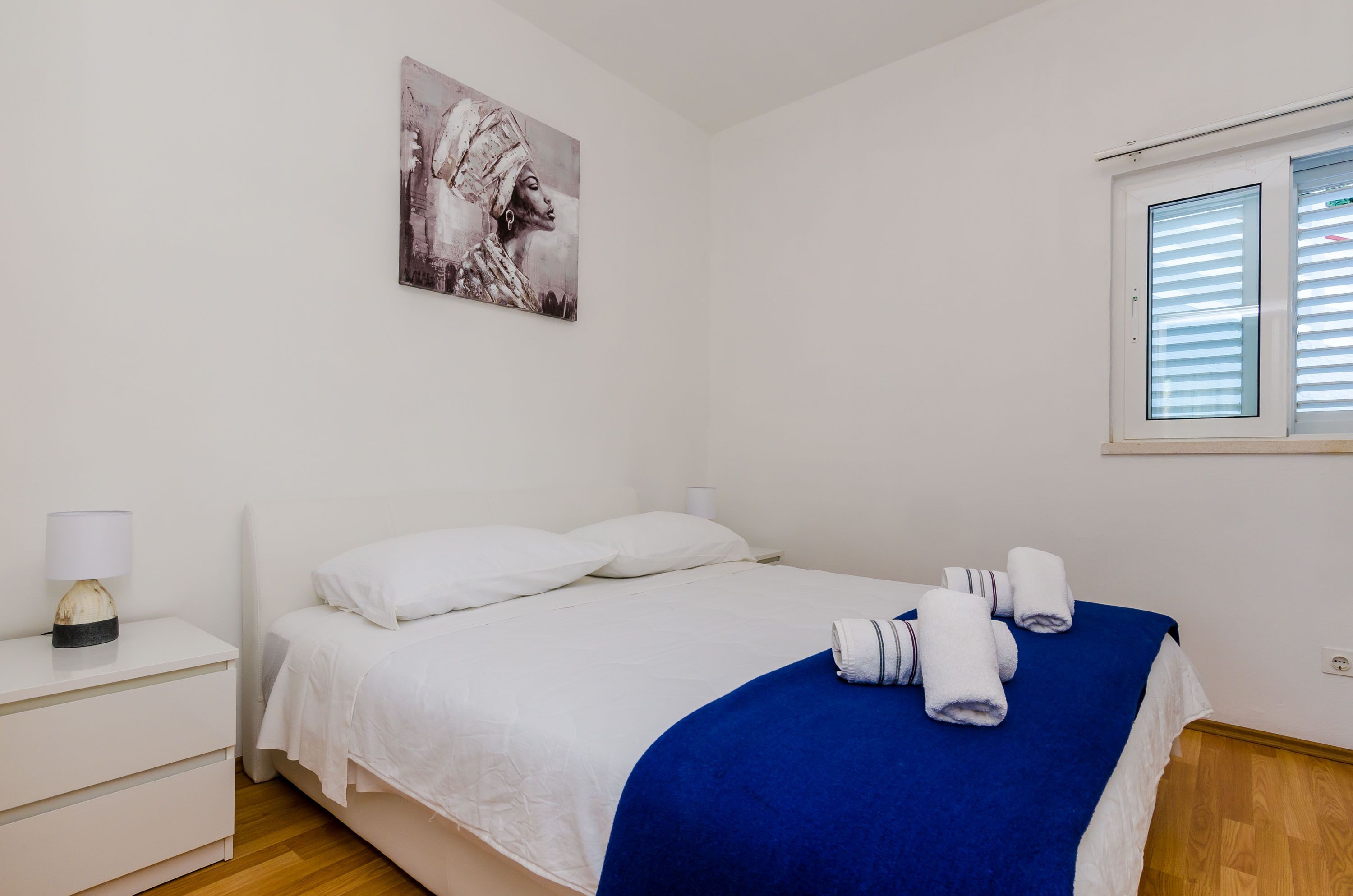 A bedroom with a bed, white nightstand, and a window with blinds.