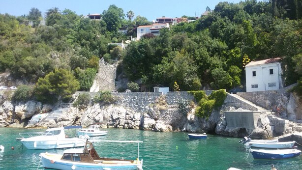 A coastal area with boats docked near a rocky shore, surrounded by lush greenery and residential buildings. - Rooms Mozara (Dubrovnik)