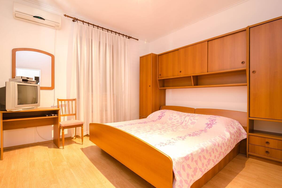 A bedroom with a bed, wooden wardrobe, a desk with a chair, and a television.