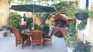 A patio with a round wooden table and chairs, a green umbrella, and a brick oven. - Rooms Mozara (Dubrovnik)