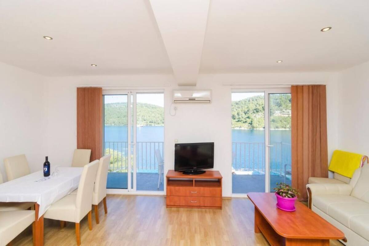 A modern living room with a dining table, a TV on a wooden stand, a sofa, and a balcony with a view of greenery.