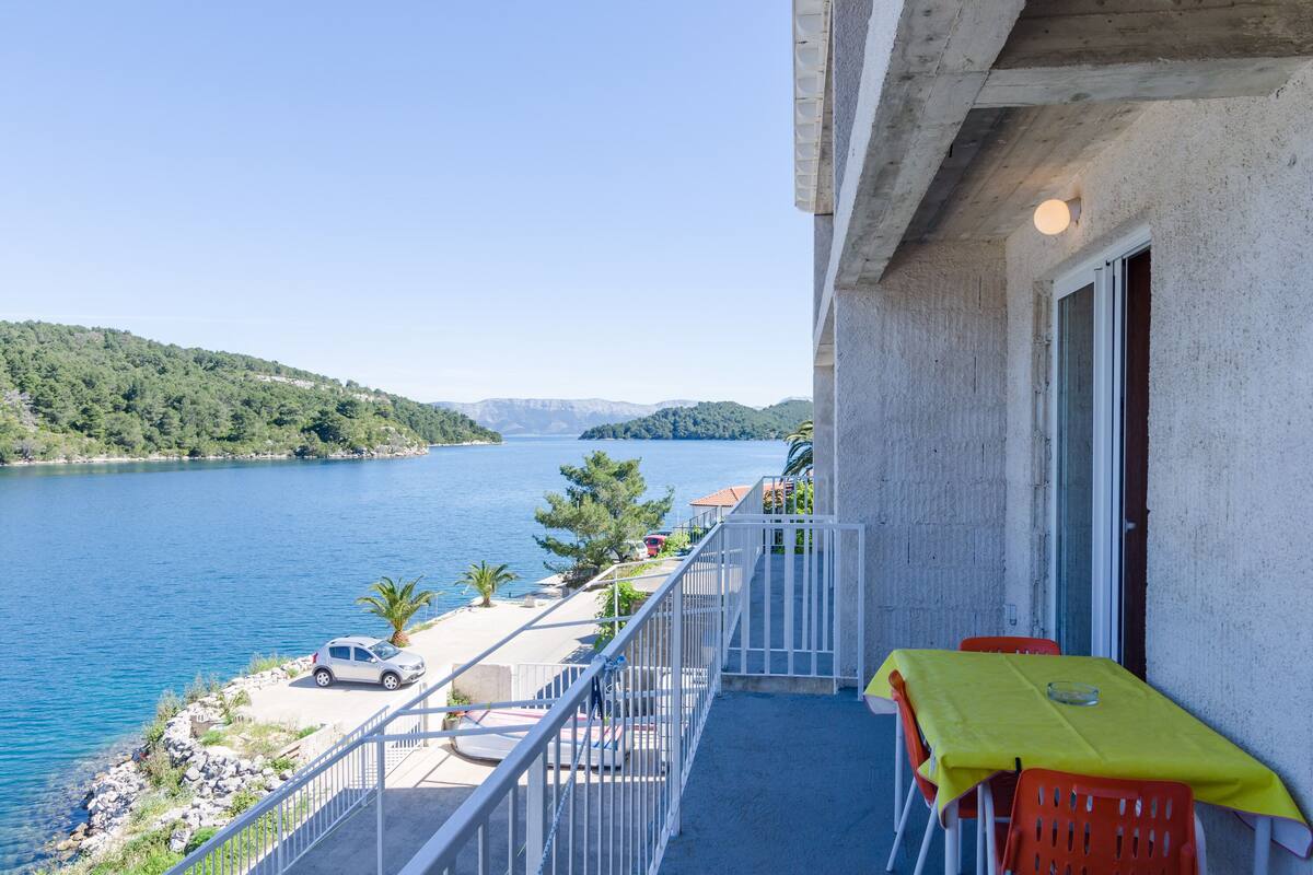 A balcony with a yellow tablecloth, a car parked by the water, and a view of the sea and hills.
