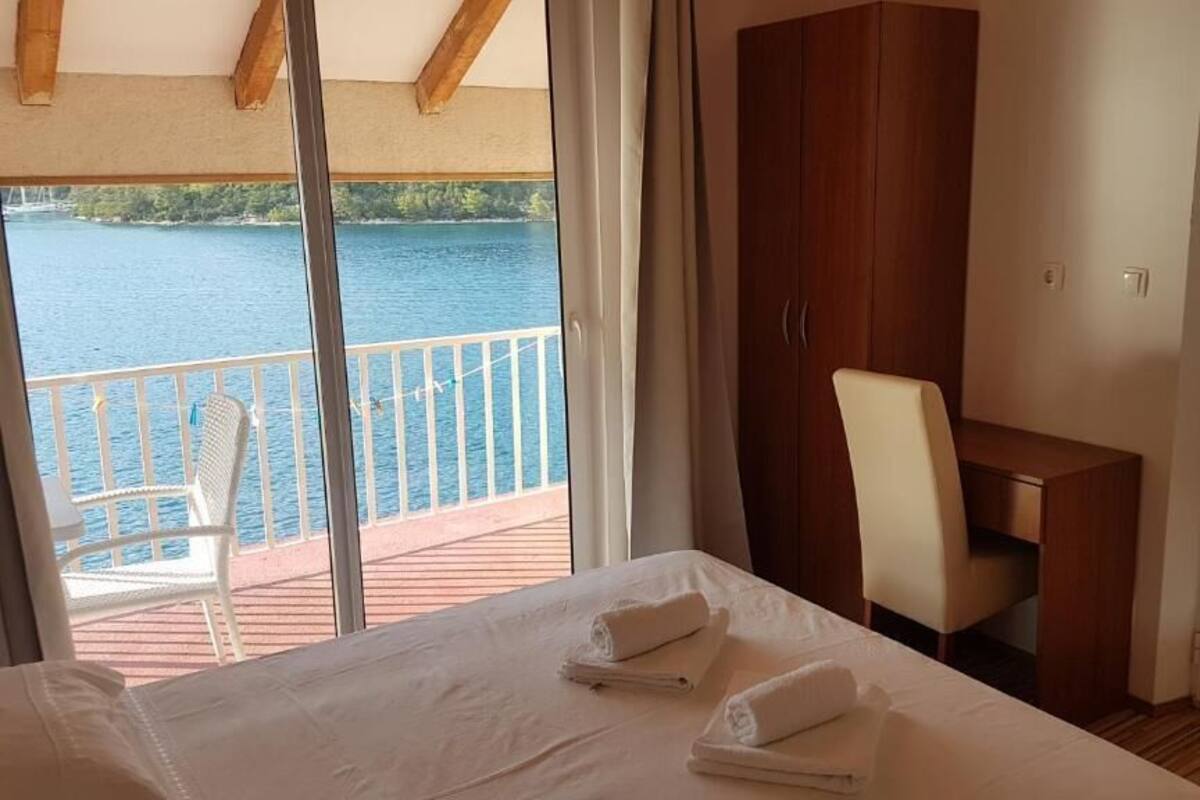 A hotel room with a bed, a desk, a chair, and a wardrobe. There is a balcony with a view of a body of water.
