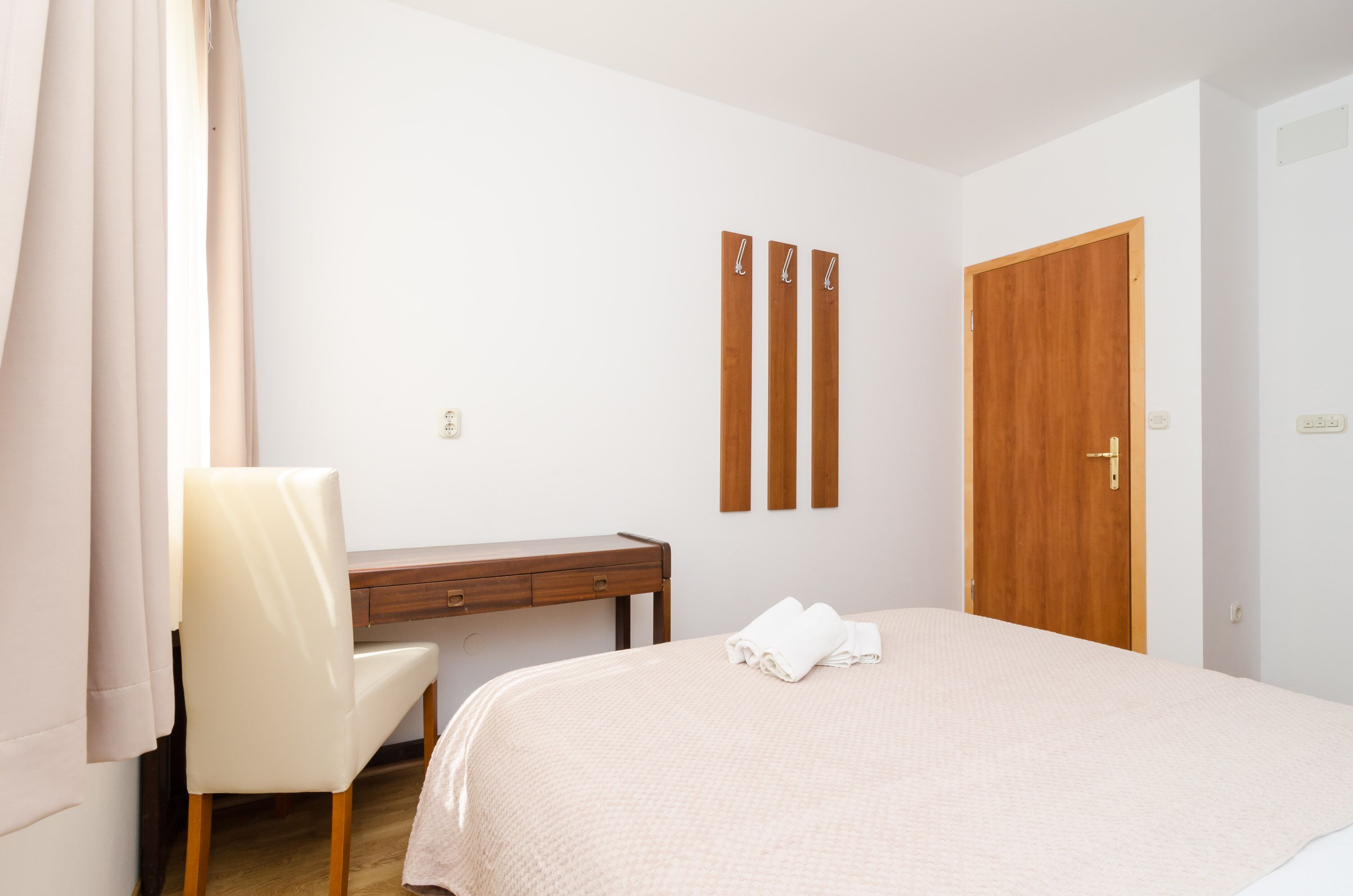 A bedroom with a bed, a desk, and a chair. There are three wooden slats on the wall and a wooden door.