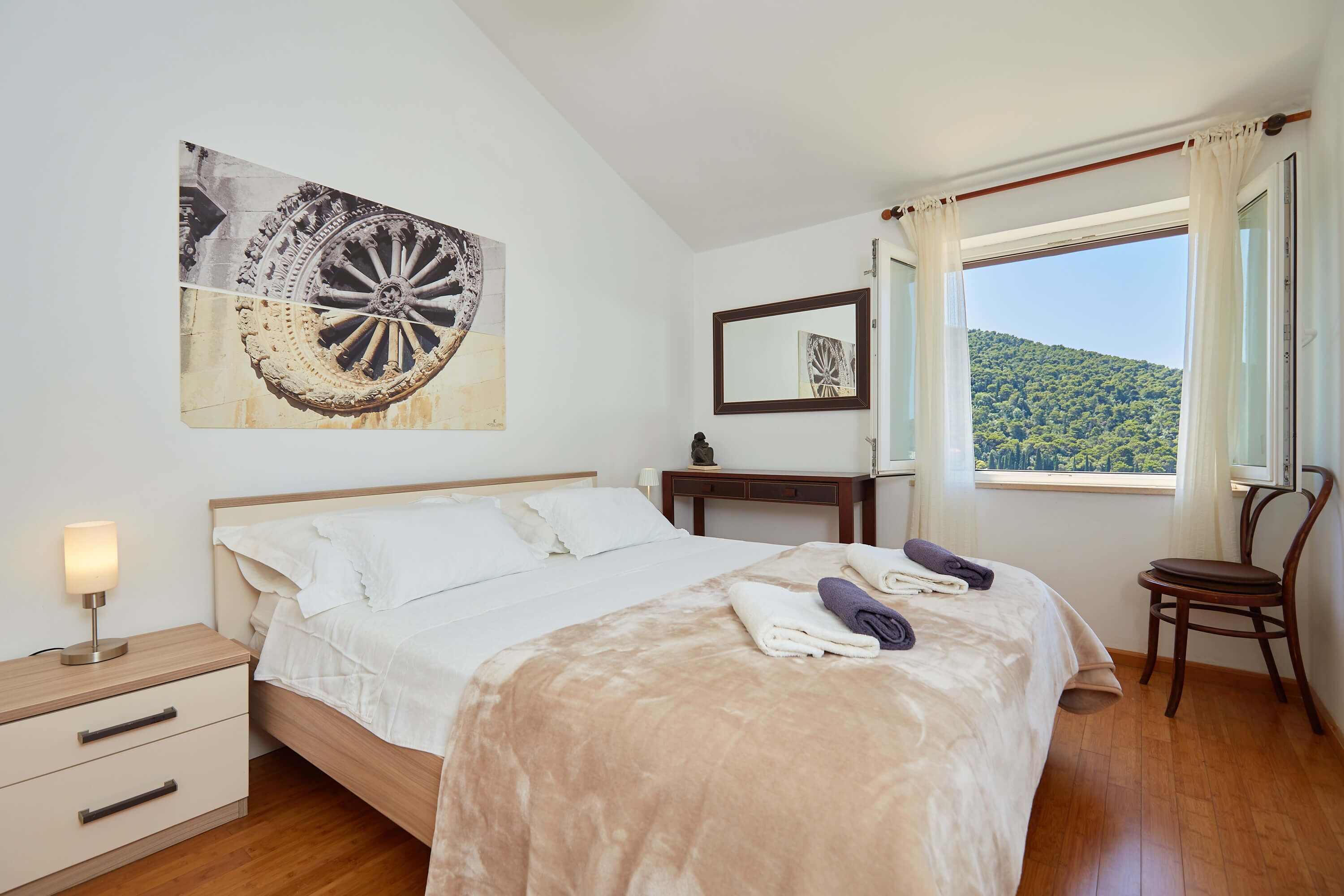 Un dormitorio con cama, mesita de noche, silla, ventana con vistas a una colina y un cuadro en la pared.