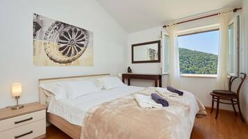 Un dormitorio con cama, mesita de noche, silla, ventana con vistas a una colina y un cuadro en la pared.