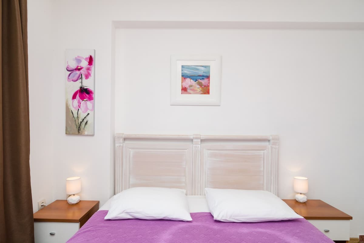 Un llit ben fet amb nínxols blancs i un edredó morat, flanquejat per dues taules de nit amb làmpades, en una habitació amb un capçal de llit blanc i parets sense adorns.