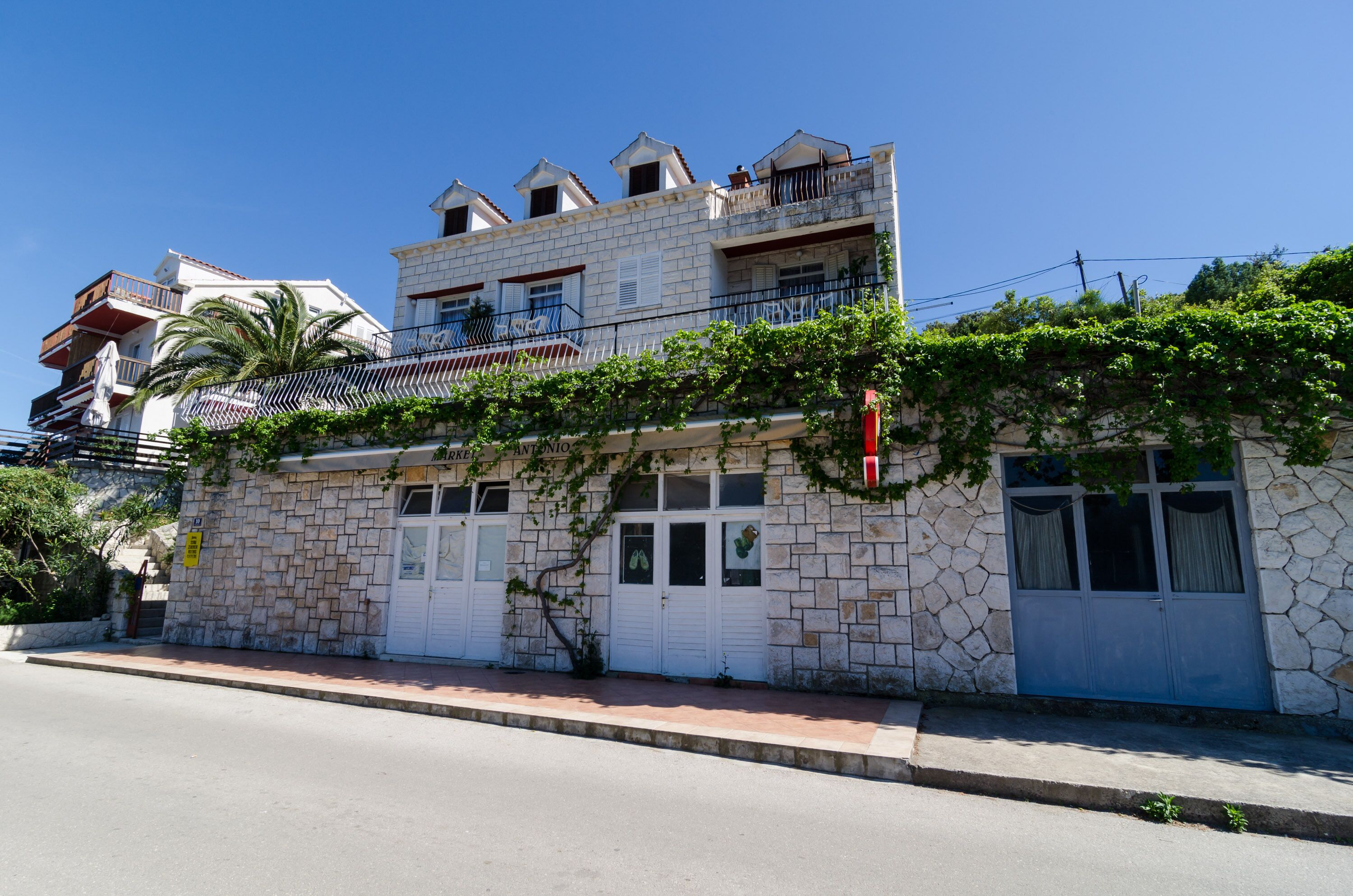A two-story stone building with a balcony, a visible door, and a small garden area.