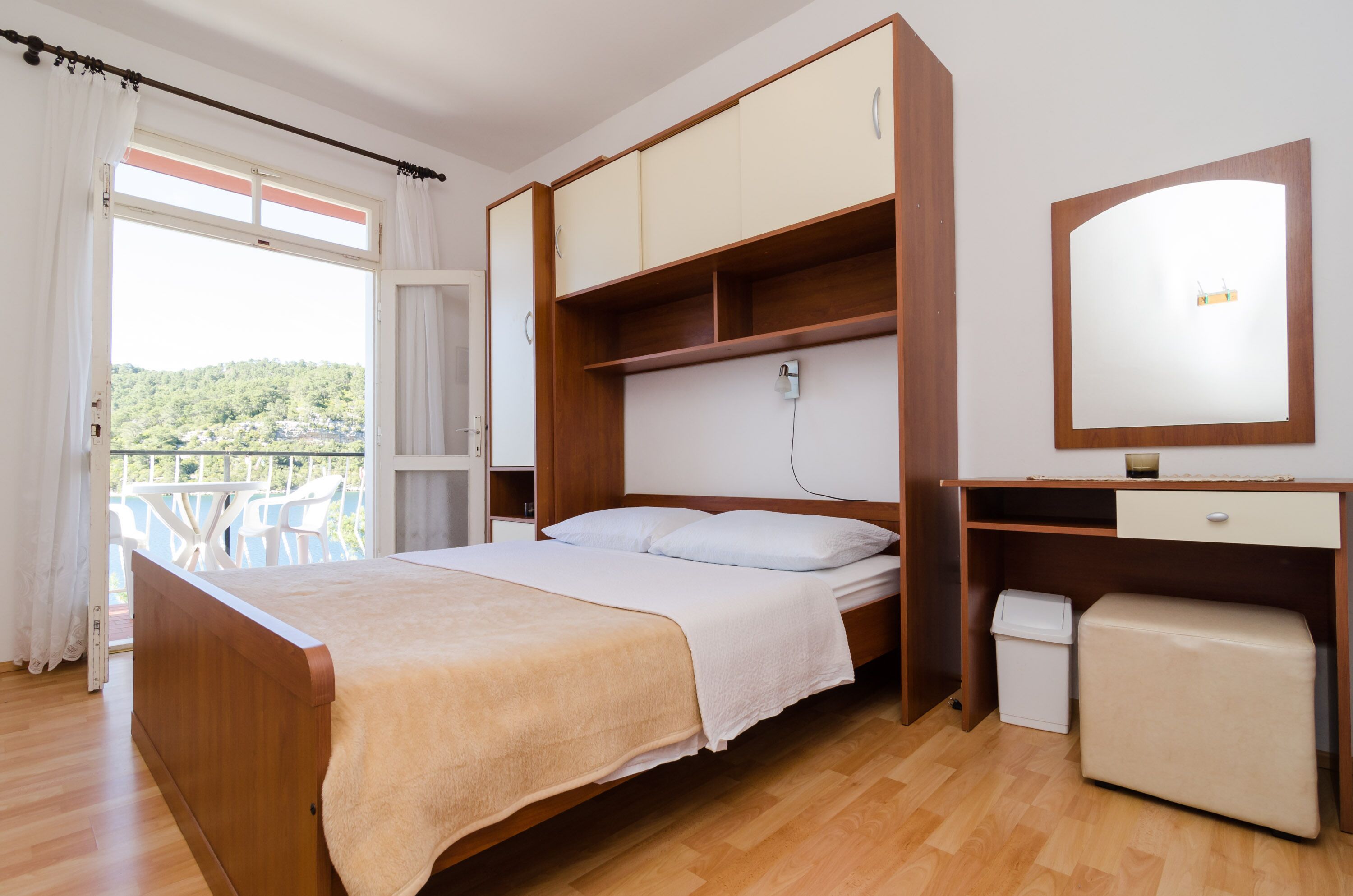 A bedroom with a bed, a wooden wardrobe, a small desk, a mirror, and a balcony with white chairs.