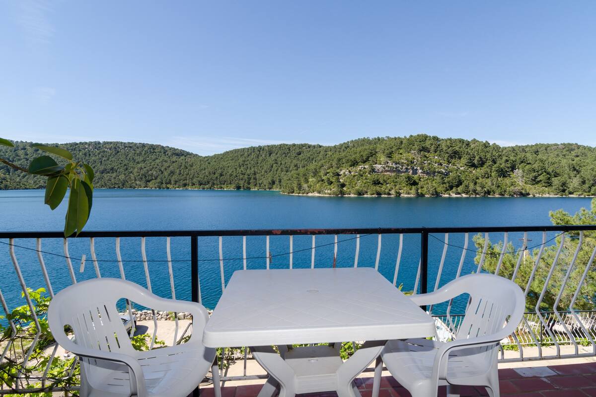 A terrace with a table and chairs overlooking a lake, surrounded by greenery and hills.