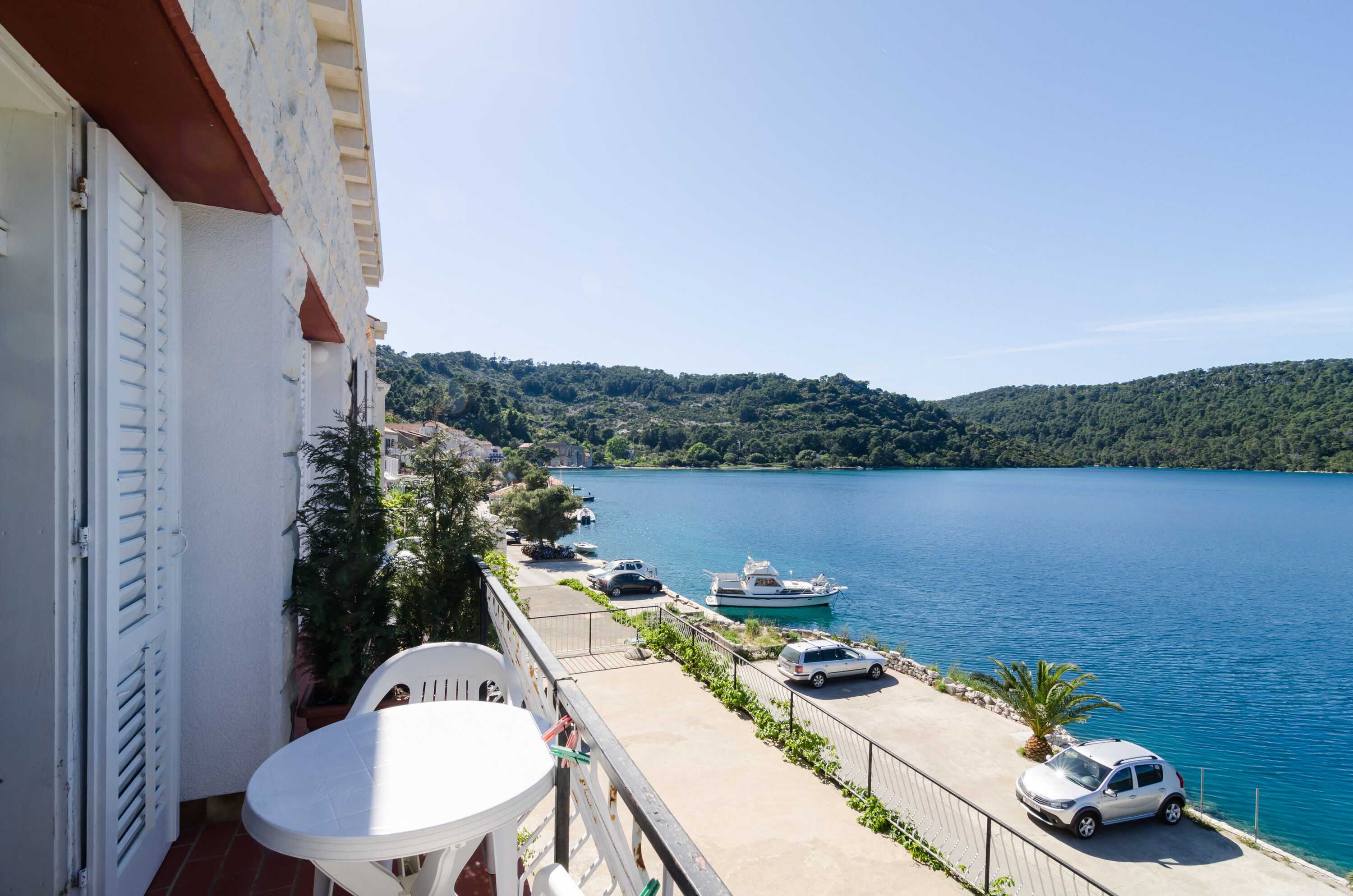 Un balcon avec une table et des chaises blanches, donnant sur un lac paisible où l’on aperçoit des bateaux et des voitures garées le long de la rive.