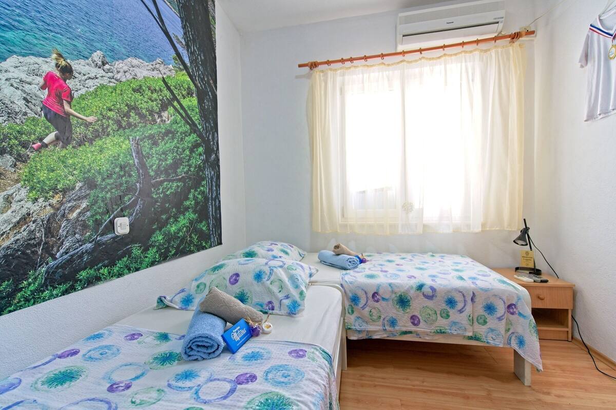 A bedroom with two beds, a window with curtains, a wall-mounted air conditioner, and a poster of a woman hiking.