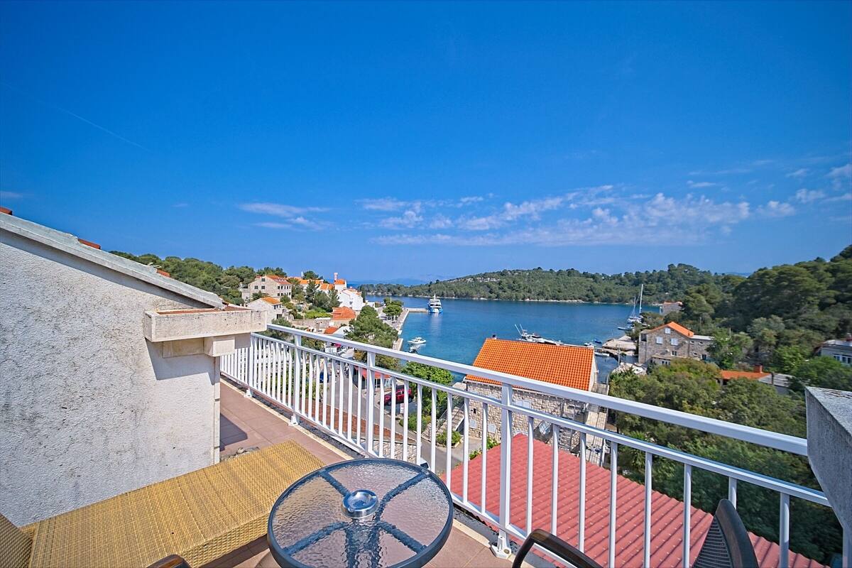 A balcony with a table and chairs, overlooking a coastal town with buildings and a body of water.