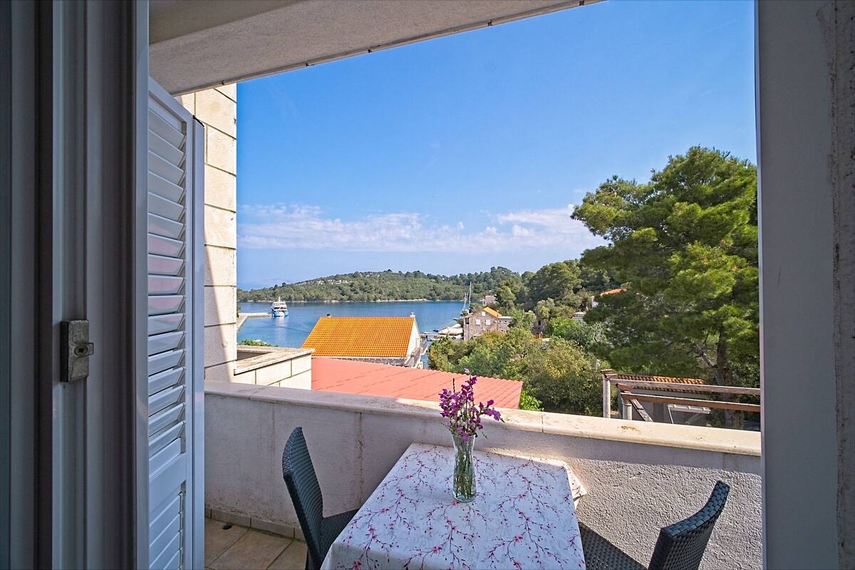 A balcony with a table and chairs, a vase with flowers, and a view of a lake and buildings.