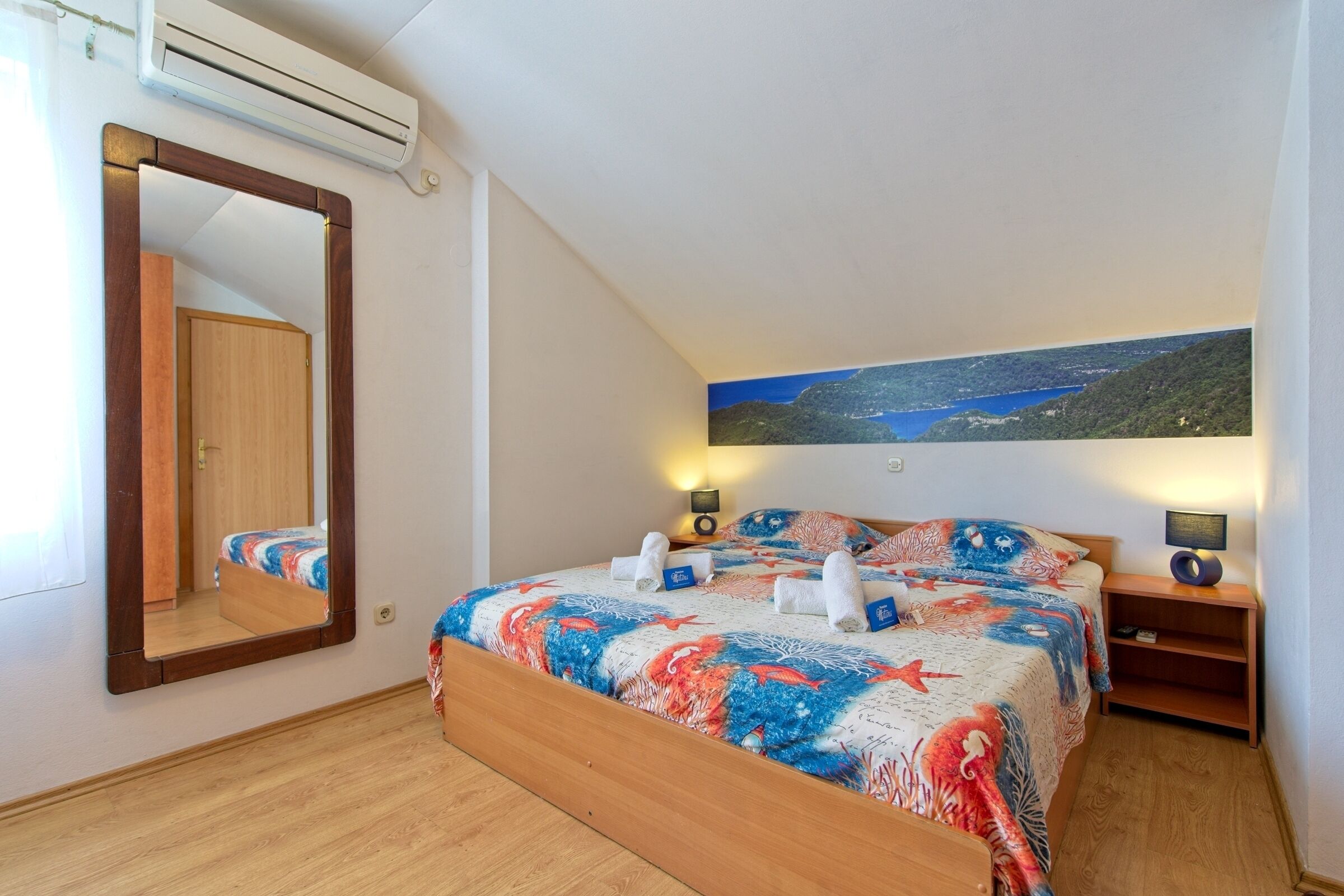 A bedroom with a bed, a large mirror, a nightstand, a wall-mounted air conditioner, and a landscape painting.