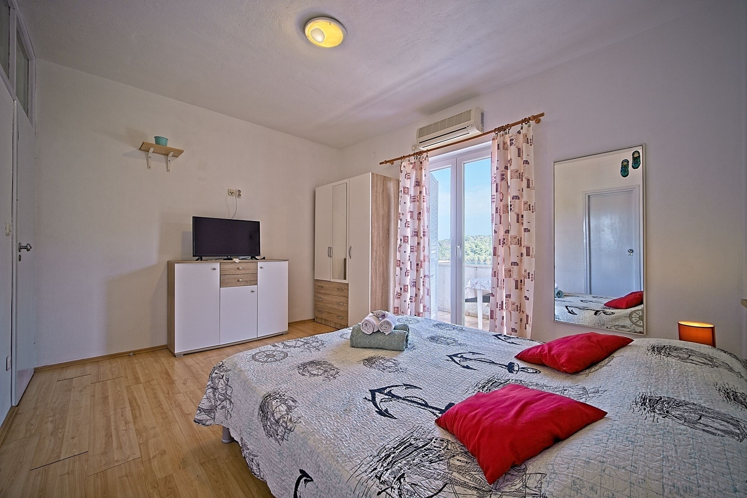 A bedroom with a bed, a TV on a stand, a wardrobe, a mirror, and a window with curtains.