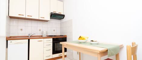 Cocina compacta con armarios blancos, lavavajillas, horno y una mesa con un bol de fruta.