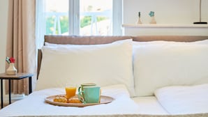 Ein ordentlich bezogenes Bett mit weißen Leinen, ein Tablett mit Croissants und Orangensaft sowie eine Lampe auf einem Nachttisch.